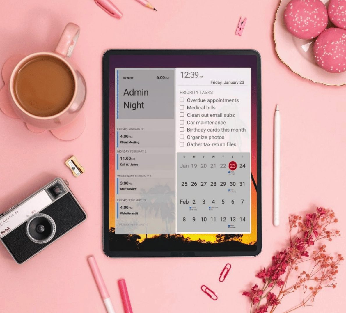 iPad on a pink desk running DAKboard with an up-next agenda, priority task list, and monthly calendar beside a camera and coffee