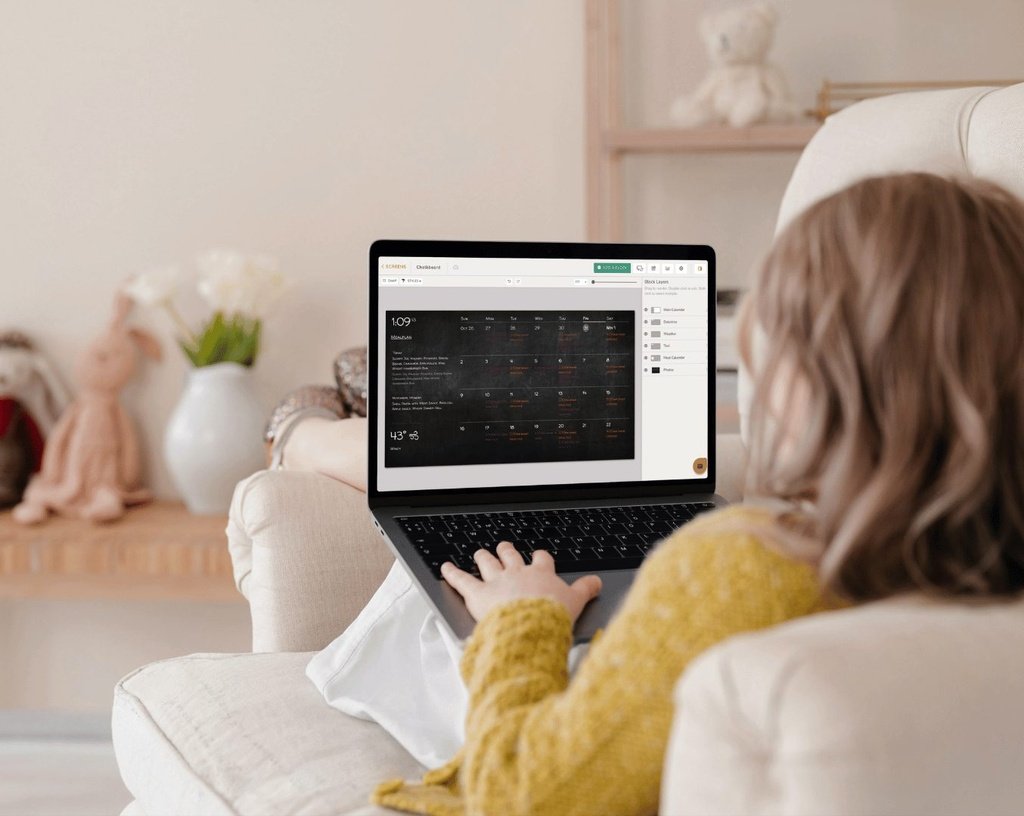 Woman on a couch using a laptop to edit a DAKboard screen layout, with the block-layers panel open beside the calendar preview