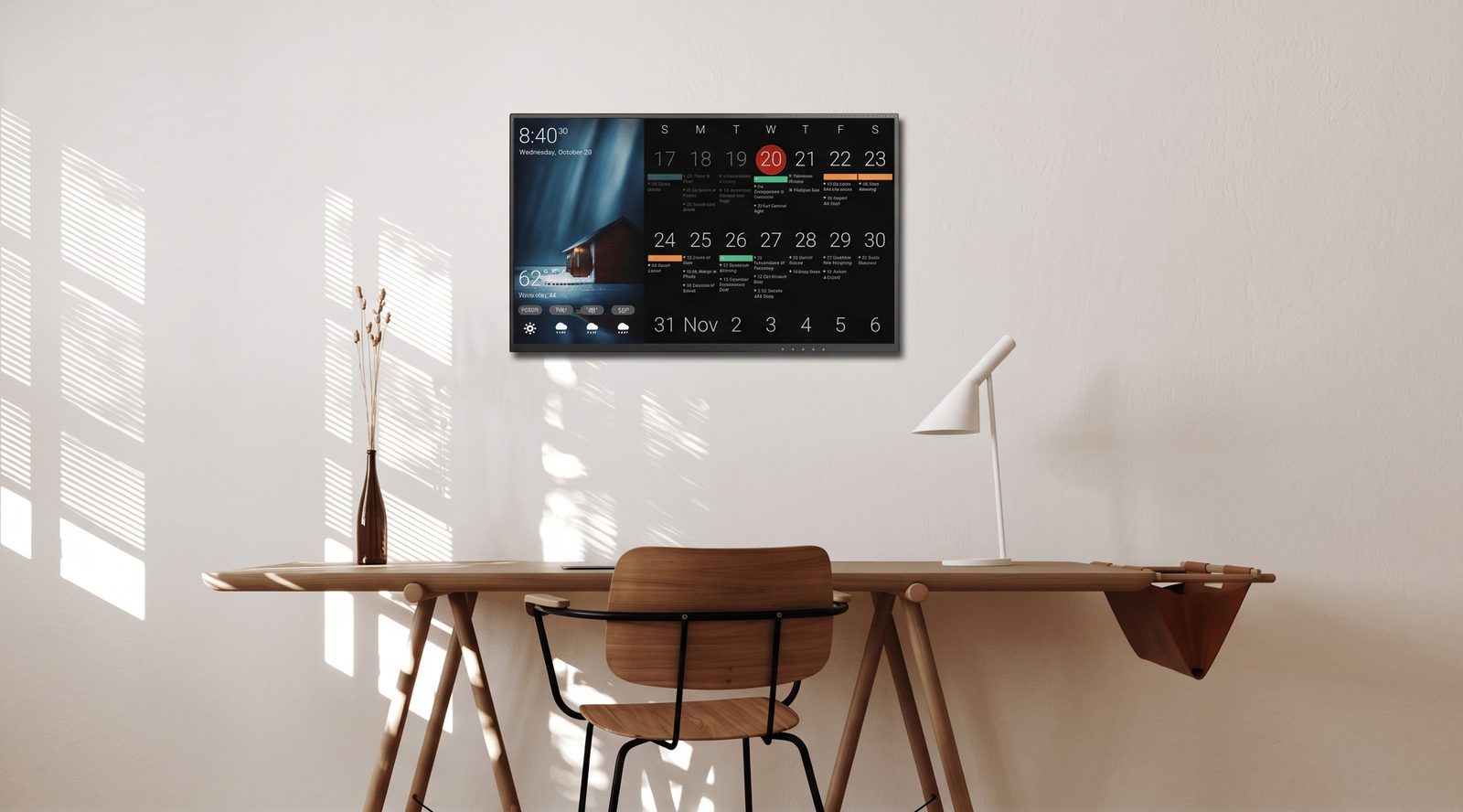 Wall-mounted DAKboard above a wooden desk showing a dark monthly calendar with color-coded events, a cabin photo, and current weather
