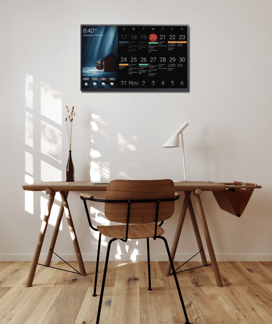 Wall-mounted widescreen display above a minimalist wooden desk shows a DAKboard monthly calendar with color-coded events and a weather panel