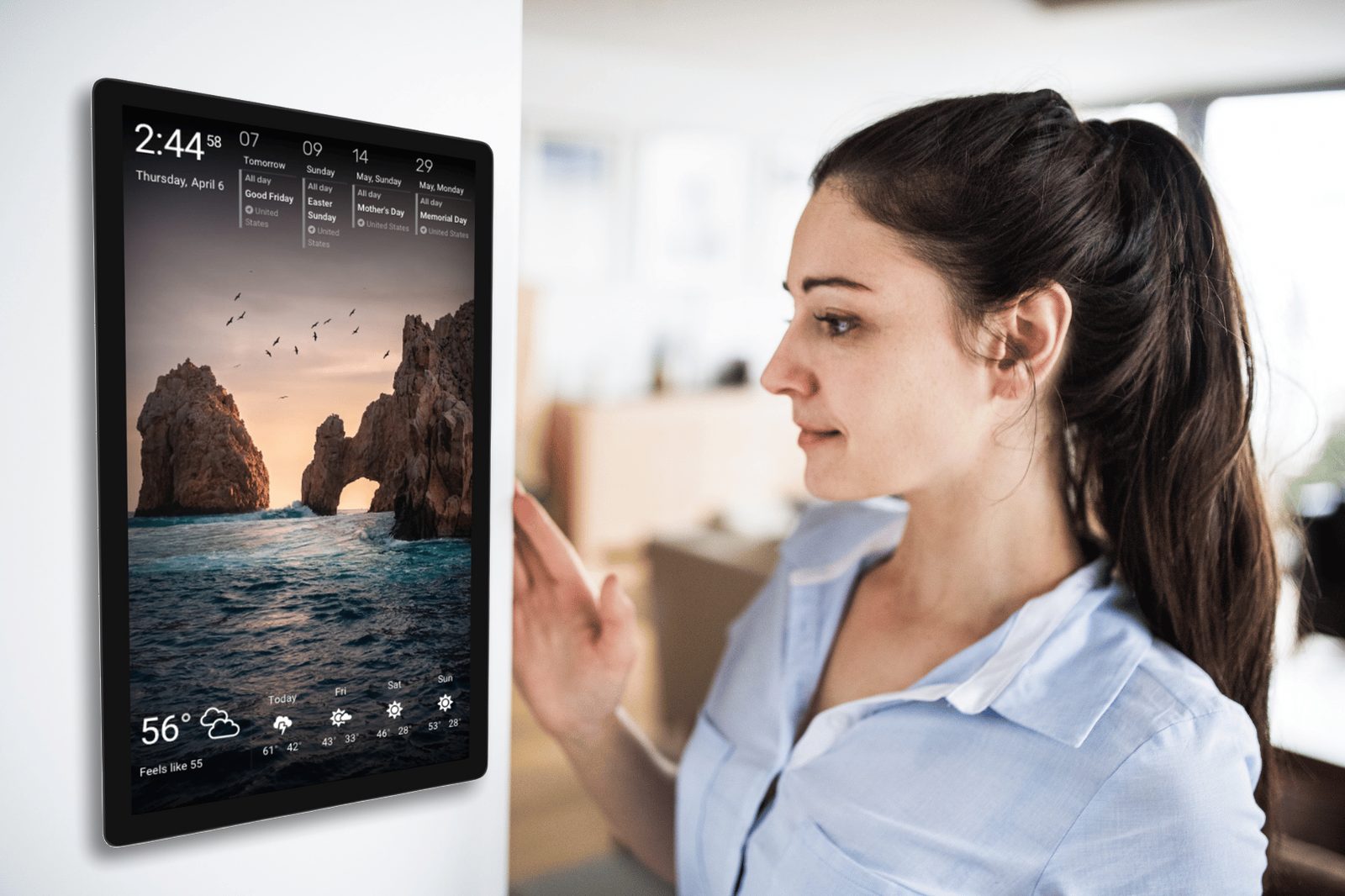 A woman glances at a wall-mounted tablet running DAKboard, showing upcoming holidays, a scenic photo, and a five-day weather forecast