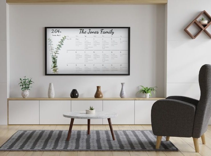 Framed family calendar display A framed DAKboard wall display in a modern living room showing a family monthly calendar titled The Jones Family
